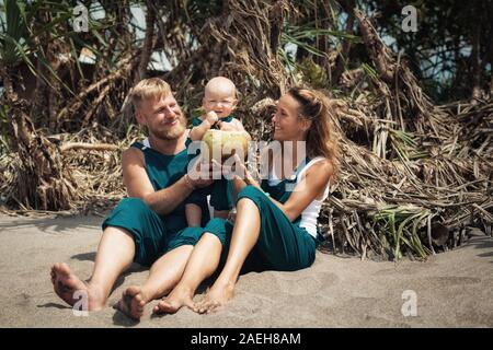 Drôle de famille heureux hipster pique-nique sur plage - mère, père nourrir bébé garçon. Petit-fils de manger des fruits avec plaisir, boire de noix de coco fraîche. Mode de vie sain. Banque D'Images