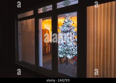 Blanc givré d'un arbre de Noël artificiel avec décoration rouge et argent vue à travers les grandes portes-fenêtres avec stores à lattes verticales et l'engrenage conique de la fenêtre Banque D'Images
