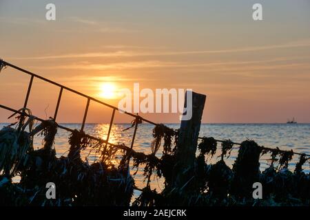 Silhouette d'une structure métallique avec des déchets plastiques et des algues en face de la mer et du coucher du soleil Banque D'Images