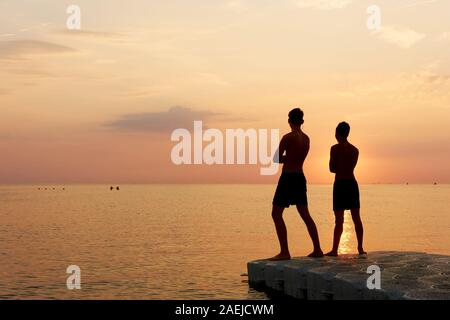 Silhouette de deux garçons en maillot de bain debout sur une jetée dans la mer face à un coucher de soleil orange with copy space Banque D'Images