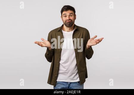 Désolés ne l'aide. Indécise et clueless beau barbu en manteau, haussant les épaules avec les mains sur le côté, sourire perplexe, ne sait rien, difficulté à Banque D'Images