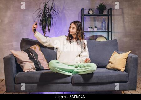 Une jeune femme assise sur un canapé dans une pièce confortable prend un sourire et selfies Banque D'Images