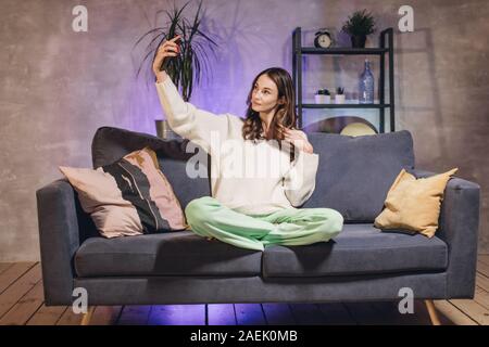 Une jeune femme assise sur un canapé dans une pièce confortable prend un sourire et selfies Banque D'Images