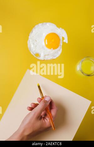 La main avec une brosse et une feuille de papier blanc tire sur le plat, à côté d'un pot d'eau jaune Banque D'Images