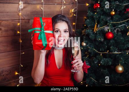 Belle femme élégante en robe rouge tenant le verre à champagne et cadeau fort près de l'arbre de Noël Banque D'Images