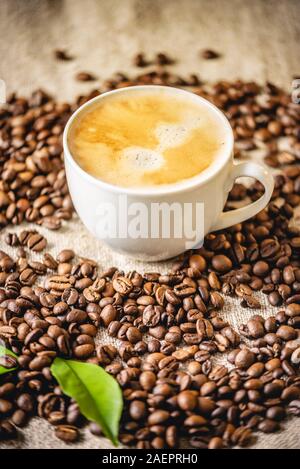 Une tasse de café avec de la mousse et des feuilles vertes sur les grains éparpillés sur la toile. Boisson revigorante parfumées à partir de grains de café fraîchement torréfié. Banque D'Images
