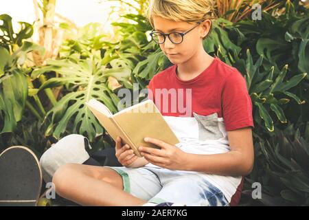 Young boy reading assis dehors dans un beau jardin tropical. Happy student faire leurs devoirs au parc enfant avec livre dans la main de détente en plein air Banque D'Images