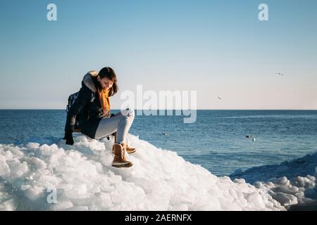 Young smiling woman sitting on a haut de la glace de mer congelés blocs sur une côte de la mer, avec un ciel bleu à l'arrière-plan, à frosty journée ensoleillée Banque D'Images