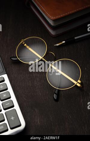 Lunettes d'or, calculatrice, bloc-notes, stylo sur un fond de bois foncé.. Banque D'Images
