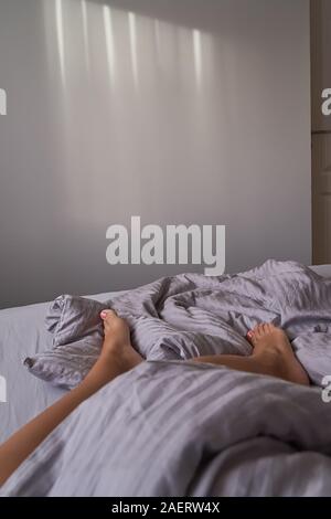 Libre les jambes des femmes sont en beige literie en chambre cosy. Matin tranquille et paresseux, concept copie espace vertical, Banque D'Images