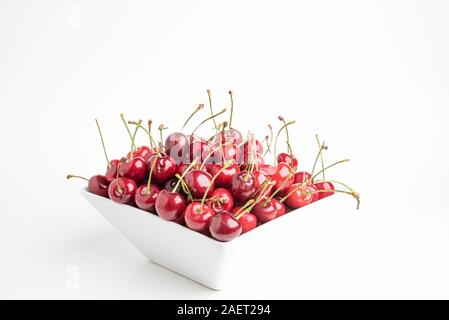 Un tas de cerises rouges frais organisé et monté dans un bol en porcelaine carré situé sur un fond blanc. Banque D'Images