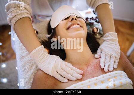 Les femmes tout en souriant thérapeute spa frottant sa poitrine. Womens mains avec des gants scrub sel rouge sur la peau de la jeune fille au centre de spa avec une décoration de Noël, Banque D'Images