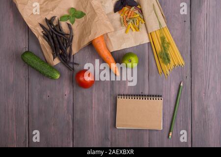 Pâtes fraîches ingrédients dans des sacs en papier écologique - pâtes spaghetti, carotte, basilic, tomates et autres légumes, le bloc-notes pour la recette sur la table en bois Banque D'Images