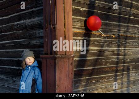 Un garçon dans un chapeau bleu oeil de derrière une clôture en bois à un ballon rouge. Banque D'Images