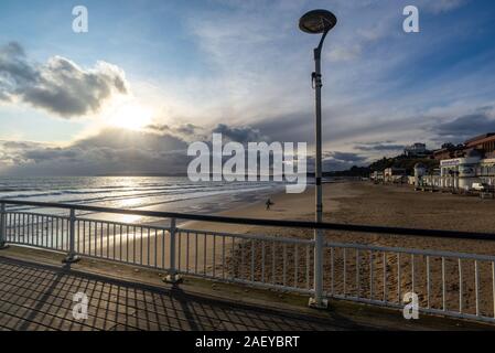 En hiver, de Bournemouth Dorset, UK, décembre 2019. Froid avec des averses de pluie, ensoleillée, avec un vent fort sur la côte sud de l'après-midi. Soleil et nuages orageux sur l'île de Purbeck. Un hardy surfer carrying un surf sur la plage après le surf dans l'eau de mer froide. Banque D'Images