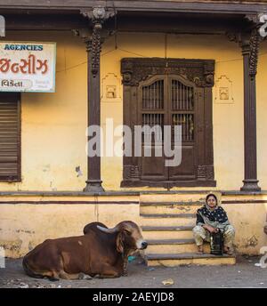 Une femme et une vache s'assoient à l'extérieur d'une vieille maison avec une porte en bois marron et des piliers en bois. Gujarati texte sur un conseil annonce que c'est une agence d'affaires. Banque D'Images