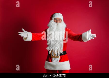L'âge moyen handsome man wearing Santa costume debout sur fond rouge isolés joyeux sourire à bras ouverts comme convivial, positif et confiant salutations. Banque D'Images