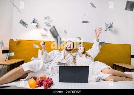 Image d'un jeune beau couple de jeunes mariés jeter de l'argent, tout en étant assis sur le lit dans la chambre d'hôtel, à l'aide d'ordinateur portable et manger des fruits. Banque D'Images