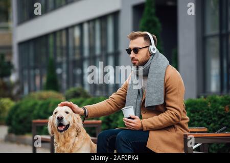 Aveugle dans les écouteurs holding thermo mug et caresser chien-guide dans park Banque D'Images