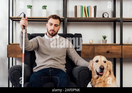 Aveugle avec bâton de marche assis dans un fauteuil à côté de golden retriever à la maison Banque D'Images