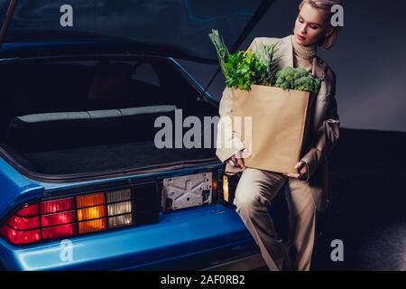 Élégante et attractive woman in suit debout près de voiture rétro et holding paper bag avec de la nourriture Banque D'Images