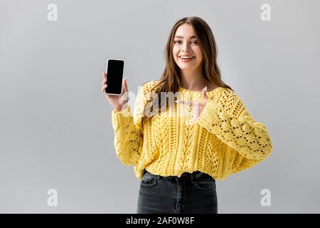 Cheerful young woman pointing avec doigt Smartphone avec écran noir sur gris isolé Banque D'Images