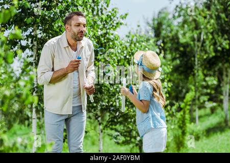 Focus sélectif de bel homme et sa fille à chapeau de paille soufflant des bulles de savon près d'arbres Banque D'Images