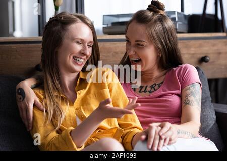 Deux jolies lesbiennes embracing while sitting on sofa in living room Banque D'Images