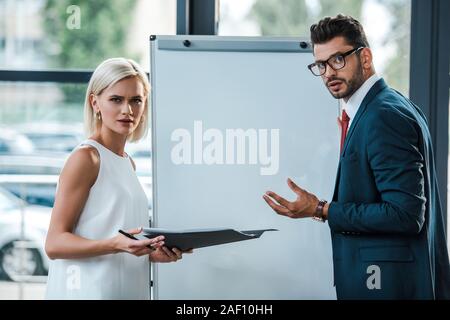 Attractive blonde woman holding folder près de businessman dans les verres des gestes in office Banque D'Images