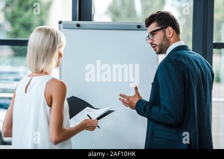 Blond woman holding folder près de businessman dans les verres des gestes in office Banque D'Images