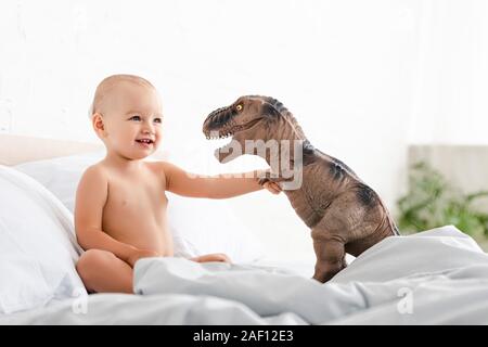 Cute little child sitting on bed, smiling and holding brown toy dinosaur Banque D'Images