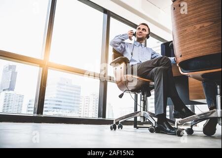 Portrait of happy handsome man with headset in modern office avec arrière-plan de construction urbaine de la ville. Businessman assis sur une chaise. Caucasian man Banque D'Images