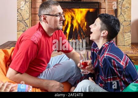 D'âge mûr de l'homme et la femme sont assis sur des sièges-sacs près de cheminée, boire du vin et le chat, ils sont heureux et joyeux. Banque D'Images