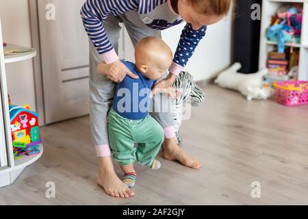 Jolie petite blonde adorable bébé garçon faire les premières étapes avec le soutien aux mères dans les jeux à la maison. Happy funny enfant qui apprend à marcher avec l'aide de MOM Banque D'Images