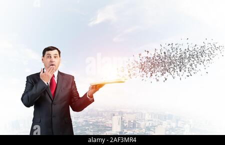 Embarrassé d'affaires couvrant la bouche avec la main et tenant un livre ouvert. L'homme d'affaires surpris en costume et cravate debout sur fond de ville avec le vol. Banque D'Images