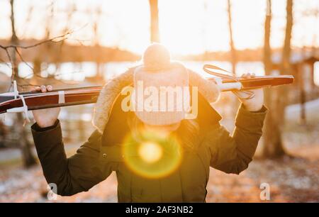 Woman carrying skis à travers une forêt enneigée au coucher du soleil Banque D'Images