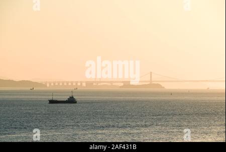 Un cargo sillonnant les rives de l'île de Naoshima, au Japon, au coucher du soleil brumeux avec le grand pont de Seto vu dans l'arrière-plan. Banque D'Images