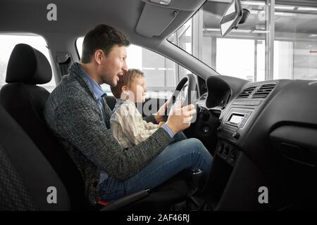 Vue du côté de funny daddy et son fils s'asseoir sur le siège de conducteurs auto, laissant les mains sur la roue et à l'avant. Machines d'essais client et le choix d'automobile. Concept de l'auto salon. Banque D'Images