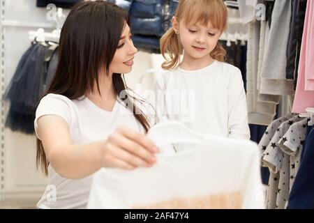 Focus sélectif de l'attractive brunette maintenant nouvelle robe dans la main et en regardant sa fille en magasin. Jeune femme le choix et l'achat des vêtements pour petite fille en boutique. Concept de l'achat. Banque D'Images