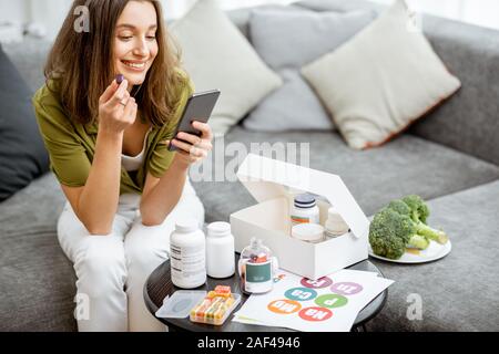 Femme de prendre des compléments alimentaires sous forme de pilules en position assise avec smart phone at home. Concept de l'individu en ligne Sélection de compléments alimentaires. La médecine préventive Banque D'Images