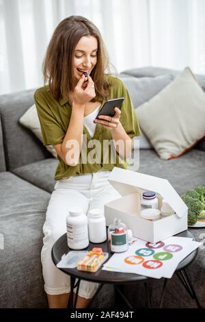 Femme de prendre des compléments alimentaires sous forme de pilules en position assise avec smart phone at home. Concept de l'individu en ligne Sélection de compléments alimentaires. La médecine préventive Banque D'Images