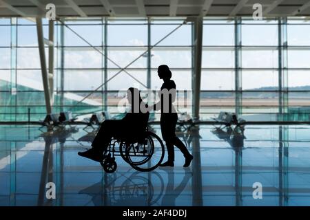 Silhouette d'une femme marchant avec une personne handicapée en fauteuil roulant à l'aéroport Banque D'Images