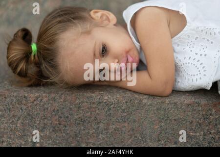 Une belle petite fille de l'apparition de cheveux blond couché sur l'asphalte de la rue triste. Les problèmes des enfants.Enfant fatigué des caprices. Banque D'Images