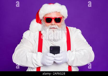 Photo de grey haired santa père holding téléphone technologie moderne de contrôle de l'utilisateur présente une usure newyear commandes sun costume violet caractéristiques isolées Banque D'Images