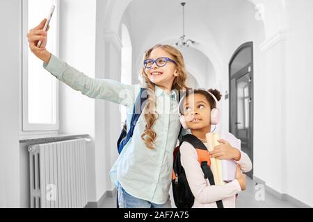 Vue de face de deux filles de l'école de friends photo smartphone et souriant, debout entre les long couloir. African and Caucasian girls avec diferent Banque D'Images