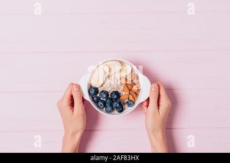 Woman's hands holding petit-déjeuner sain de gruau avec des amandes, les bleuets et les bananes dans un bol blanc rose sur fond de bois, vue du dessus Banque D'Images