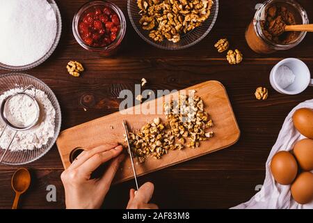 Top View woman's hands hacher les noix pour la cuisson du gâteau brownie parmi les ingrédients sur la table en bois sombre. Cuisine maison, processus de mise à plat. Banque D'Images