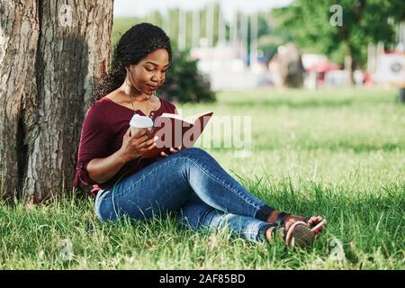 Lecture et est titulaire d'un verre dans la main. Cheerful african american woman dans le parc à l'été Banque D'Images