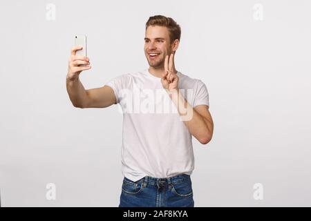 Cheerful man télécharger nouvelle app, en essayant de prendre comme filtres mignon sur smartphone selfies, faisant signe de la paix et souriant joyeusement, photographier lui-même Banque D'Images