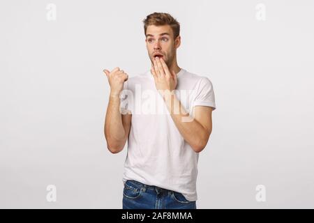 Surpris et choqué jeune blonde beau mec, ponting pouce gauche et insistants, de halètement couvrir la bouche comme le commérage, témoin de quelque chose de surprenant Banque D'Images